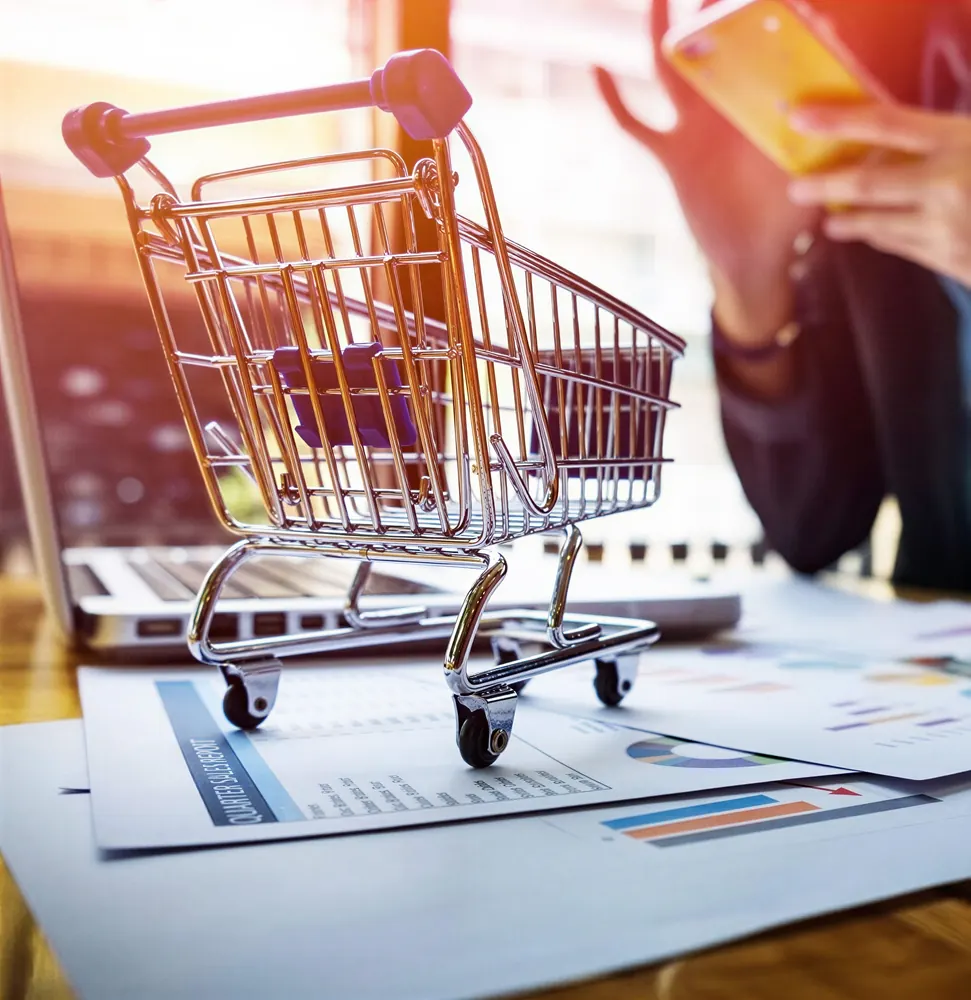 Mini carrinho de compras sobre relatórios de vendas ao lado de um notebook, representando o acompanhamento financeiro mensal do e-commerce