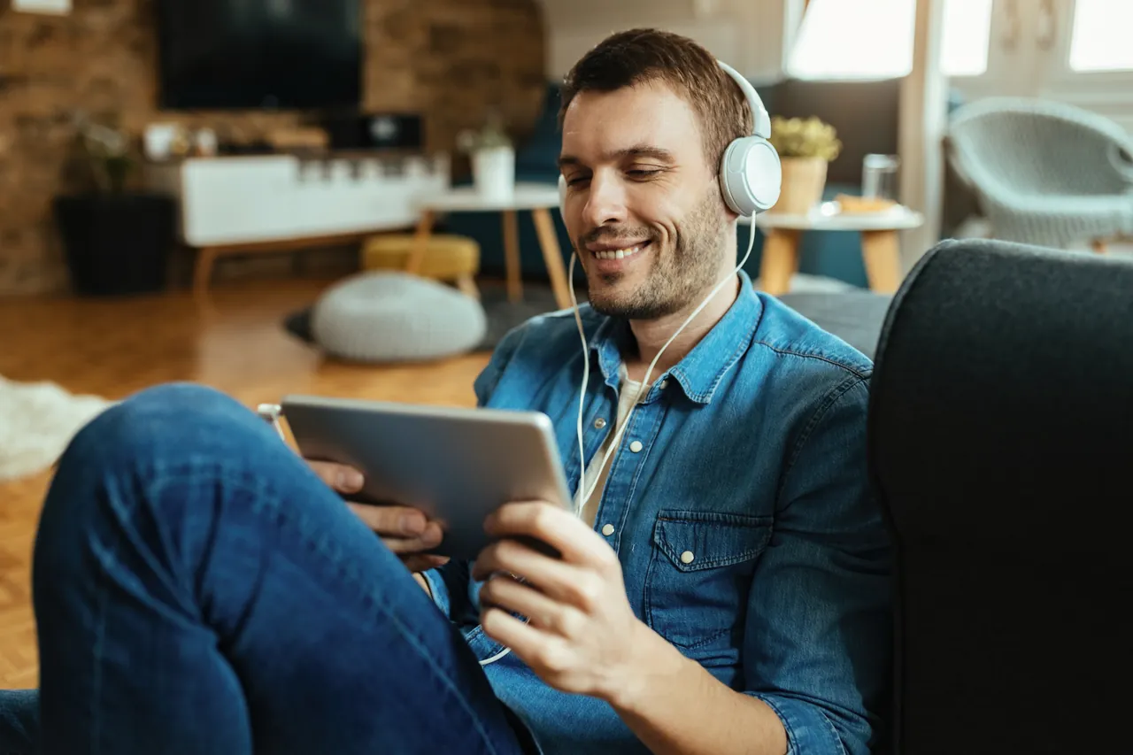 Homem sorridente no sofá, ouvindo música e consultando um tablet, representando a tranquilidade de acompanhar os números do mês